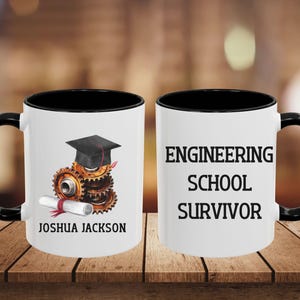 Könnte beinhalten: Weißer Keramikbecher mit schwarzem Innenraum und Henkel. Eine Seite zeigt eine Abschlusskappe auf Zahnrädern und einer Schriftrolle mit dem Namen "JOSHUA JACKSON". Die andere Seite lautet "ENGINEERING SCHOOL SURVIVOR."