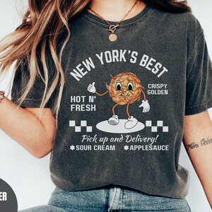Pu&ograve; includere: Una maglietta grigia scura con un'immagine di una patata di cartone animato con il testo "New York's Best Hot N' Fresh Crispy Golden" e "Pick up and Delivery! Sour Cream Applesauce".
