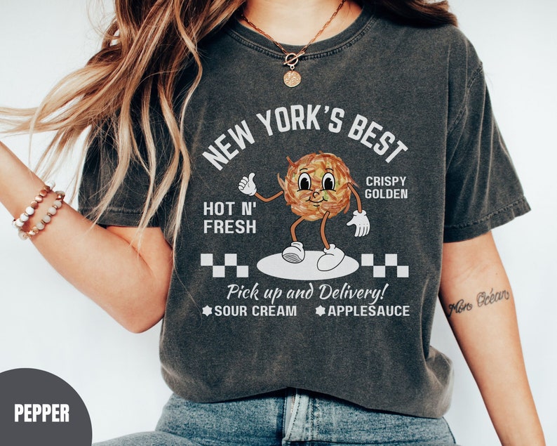 Pu&ograve; includere: Una maglietta grigia scura con un'immagine di una patata di cartone animato con il testo "New York's Best Hot N' Fresh Crispy Golden" e "Pick up and Delivery! Sour Cream Applesauce".