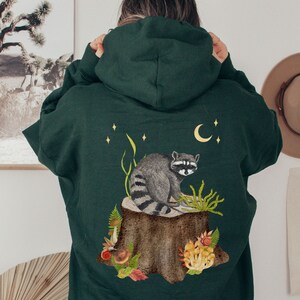 May include: A dark green hooded sweatshirt with a raccoon sitting on a tree stump surrounded by mushrooms and leaves. The raccoon is gray and black with a white mask. The design is on the back of the sweatshirt.