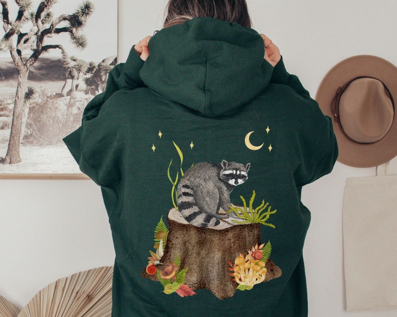 May include: A dark green hooded sweatshirt with a raccoon sitting on a tree stump surrounded by mushrooms and leaves. The raccoon is gray and black with a white mask. The design is on the back of the sweatshirt.