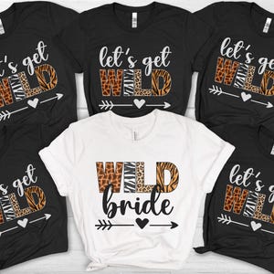 May include: Five short-sleeved t-shirts arranged in a group. Four black shirts feature the white text "let's get" above the word "WILD" in animal print patterns, with a heart and arrow. One white shirt features "WILD" in animal print and "bride" in black.
