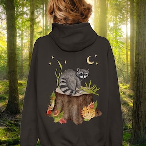 May include: A dark gray hoodie with a raccoon sitting on a tree stump surrounded by mushrooms and leaves. The raccoon is looking to the right. There are stars and a crescent moon in the background.