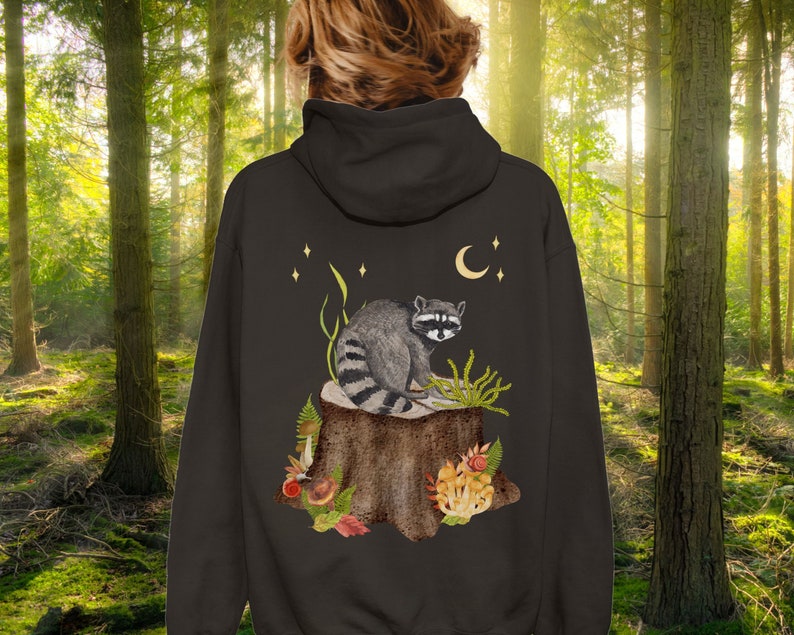 May include: A dark gray hoodie with a raccoon sitting on a tree stump surrounded by mushrooms and leaves. The raccoon is looking to the right. There are stars and a crescent moon in the background.