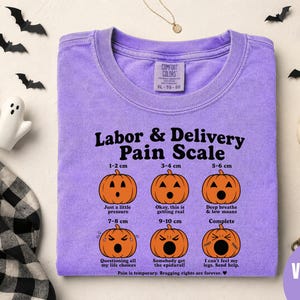 May include: A violet t-shirt featuring a black graphic titled Labor & Delivery Pain Scale. The design displays six orange jack-o'-lanterns with varying facial expressions, labeled with measurements from 1-2 cm to 10 cm and descriptive text.