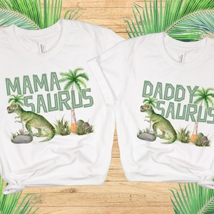 May include: White t-shirts with green dinosaur and palm tree graphics. The text on the shirts reads "Mama Saurus" and "Daddy Saurus".