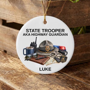 May include: A round white ceramic ornament featuring the text STATE TROOPER AKA HIGHWAY GUARDIAN and LUKE. The graphic includes a campaign hat, a blue mug, a radio, sunglasses, a pen, a notebook, handcuffs, a flashlight, keys, and a badge.