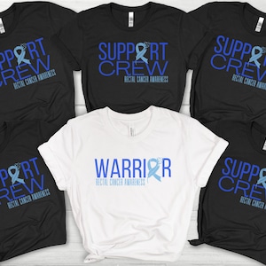 Puede incluir: Seis camisetas con diseños de cinta azul. Cinco camisetas negras con el texto "Support Crew" y una cinta azul. Una camiseta blanca con el texto "Warrior" y una cinta azul. Todas las camisetas tienen el texto "Concientización sobre el cáncer rectal".