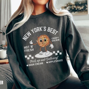 Pu&ograve; includere: Una felpa grigia scura con un design grafico bianco che dice "New York's Best" con un'immagine di un cibo croccante e dorato in stile cartone animato. Il testo dice anche "Hot N' Fresh", "Crispy Golden", "Pick up and Delivery!" e "Sour Cream Applesauce".
