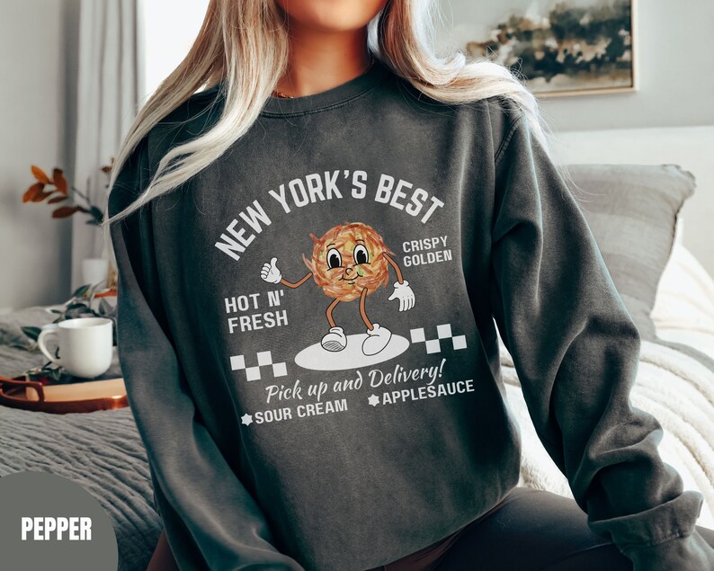 Pu&ograve; includere: Una felpa grigia scura con un design grafico bianco che dice "New York's Best" con un'immagine di un cibo croccante e dorato in stile cartone animato. Il testo dice anche "Hot N' Fresh", "Crispy Golden", "Pick up and Delivery!" e "Sour Cream Applesauce".