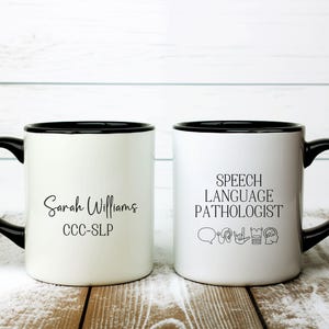 May include: Two white ceramic mugs with black handles and rims. One mug features the name "Sarah Williams" and "CCC-SLP" in black script. The other mug displays "SPEECH LANGUAGE PATHOLOGIST" with related icons.