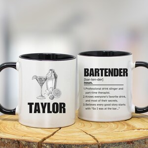 May include: Two white ceramic mugs with black handles and rims. One mug features a cocktail illustration and the name "TAYLOR". The other mug has the word "BARTENDER" and a definition.