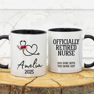 May include: Two white ceramic mugs with black handles and rims. One mug features a stethoscope design with floral accents and the name "Amelia" and the year "2025". The other mug reads "OFFICIALLY RETIRED NURSE" and "100% DONE WITH THIS WORK SHIT".