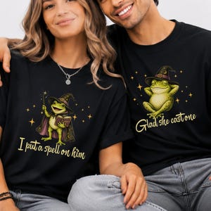 May include: Two black t-shirts featuring green frog illustrations. The left shirt shows a frog in a witch hat holding a book and wand with the text I put a spell on him. The right shirt shows a frog in a witch hat with the text Glad she cast one.