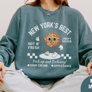 Pu&ograve; includere: Una felpa blu con una grafica di un anello di cipolla di cartone animato con il testo "New York's Best Crispy Golden Hot N' Fresh Pick up and Delivery! Sour Cream Applesauce" stampato sul davanti. La felpa &egrave; di colore blu abete.