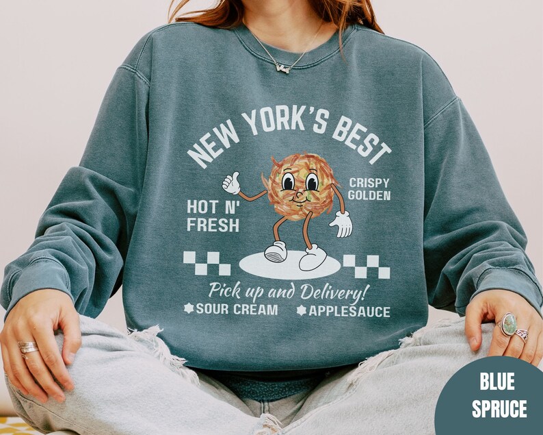Pu&ograve; includere: Una felpa blu con una grafica di un anello di cipolla di cartone animato con il testo "New York's Best Crispy Golden Hot N' Fresh Pick up and Delivery! Sour Cream Applesauce" stampato sul davanti. La felpa &egrave; di colore blu abete.