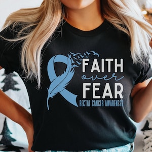 Puede incluir: Camiseta negra con una cinta azul, una pluma y el texto "FAITH over FEAR" con las palabras "RECTAL CANCER AWARENESS" debajo.