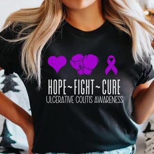 May include: Black t-shirt with a purple heart, boxing gloves, and ribbon. The text "HOPE - FIGHT - CURE" and "ULCERATIVE COLITIS AWARENESS" is printed on the shirt.