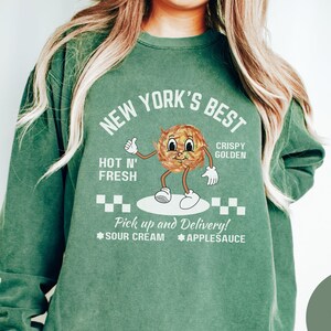 Pu&ograve; includere: Una felpa verde chiaro con il testo "NEW YORK'S BEST" in bianco. La felpa presenta un'illustrazione di un cibo dorato e croccante con un viso sorridente. Il testo "HOT N' FRESH" e "CRISPY GOLDEN" &egrave; anche stampato sulla felpa. Il testo "Pick up and Delivery!" &egrave; stampato sotto l'illustrazione, insieme alle parole "SOUR CREAM" e "APPLESAUCE".