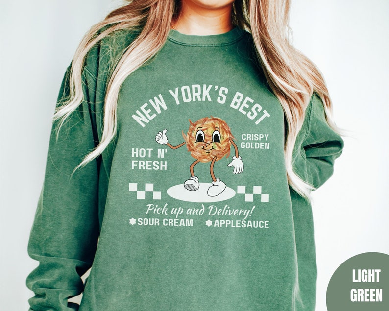 Pu&ograve; includere: Una felpa verde chiaro con il testo "NEW YORK'S BEST" in bianco. La felpa presenta un'illustrazione di un cibo dorato e croccante con un viso sorridente. Il testo "HOT N' FRESH" e "CRISPY GOLDEN" &egrave; anche stampato sulla felpa. Il testo "Pick up and Delivery!" &egrave; stampato sotto l'illustrazione, insieme alle parole "SOUR CREAM" e "APPLESAUCE".
