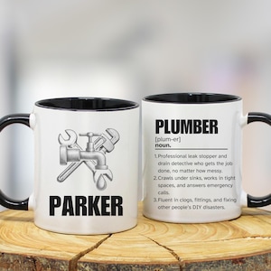 Puede incluir: Dos tazas de cerámica blanca con asas y bordes negros. Una taza presenta un gráfico de grifo y llave inglesa con el nombre "PARKER". La otra taza tiene la palabra "PLUMBER" y una definición.