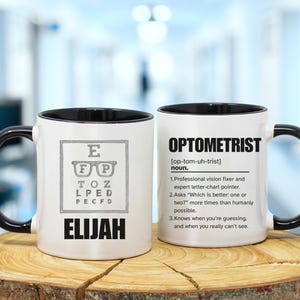 May include: White ceramic mug with a black handle and rim. One side features an eye chart and the name "ELIJAH" in bold black letters. The other side has the word "OPTOMETRIST" and its definition.