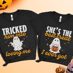May include: Two black t-shirts featuring white ghost graphics and orange text. The left shirt reads TRICKED him into loving me. The right shirt reads SHE'S THE best treat I ever got. Both shirts include small star accents and ghost illustrations.