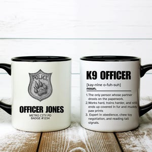 Könnte beinhalten: Weißer Keramikbecher mit schwarzem Henkel und Rand. Eine Seite zeigt ein Polizeischild mit Pfotenabdrücken und dem Text "OFFICER JONES". Die andere Seite hat "K9 OFFICER" und eine Definition. Ein tolles Geschenk.