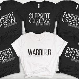 Puede incluir: Cinco camisetas con un tema de concienciación sobre el cáncer cerebral. Cuatro camisetas negras con el texto "Support Crew" y una cinta gris. Una camiseta blanca con el texto "Warrior" y una cinta gris.