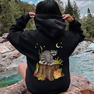May include: Black hooded sweatshirt with a graphic print of a raccoon sitting on a tree stump with a crescent moon and stars in the background.