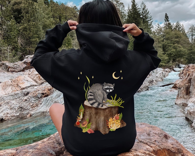 May include: Black hooded sweatshirt with a graphic print of a raccoon sitting on a tree stump with a crescent moon and stars in the background.