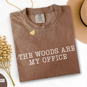 May include: Espresso-colored t-shirt with the text "THE WOODS ARE MY OFFICE" in white. A gold heart pendant necklace and a beige hat are next to the shirt. A bouquet of dried flowers is also in the image.