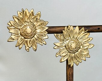 May include: A pair of gold-tone sunflower earrings. The earrings are displayed on a wooden stand.