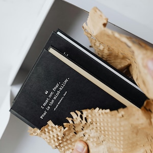 Può includere: Due diari con copertina rigida neri con una citazione bianca sulla copertina: "Non devo temere. La paura è l'assassino della mente." -Frank Herbert, Dune. I diari sono avvolti in carta da imballaggio marrone.