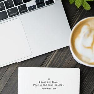 May include: A white laptop computer with a black keyboard and a white trackpad is on a wooden table. A cup of coffee with a white foam design is on the table. A white card with a quote from Frank Herbert's Dune is on the table. The quote reads "I must not fear. Fear is the mind-killer."