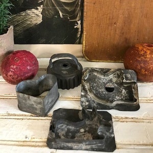 May include: An assortment of vintage metal cookie cutters in various shapes, including a heart, a circle, and a horse. A sepia-toned photograph of a woman and child is in the background, along with a wooden cutting board and a red decorative ball.