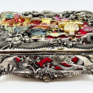 May include: A rectangular metal box featuring intricate silver-toned dragon reliefs and a central scene with gold, red, and blue accents. The lid and sides display detailed mythical creatures and architectural motifs, supported by four ornate clawed feet.