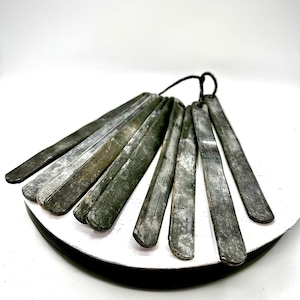 May include: A set of seven elongated, flat, dark gray metal strips with a weathered, distressed finish. Each strip features a rounded end and a small hole at the top, all connected together by a thin black cord, creating a fan-like arrangement.