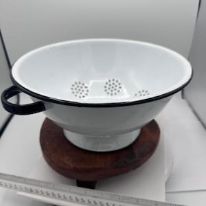 May include: White enamel colander with black trim and handle. The colander has small drainage holes and sits on a wooden base. A black ruler is in the foreground.