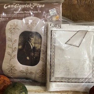 May include: A decorative picture frame kit with a photo inside, and a second kit with a needlework design. The frame is white with floral and snowflake embellishments. The photo is of a person in a dark suit. The frame finishes 10" x 12".