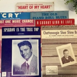 May include: A collection of vintage sheet music covers, featuring titles like "Heart of My Heart," "Cry," and "Chattanoogie Shoe Shine Boy." The covers display various colors and include images of singers and text.
