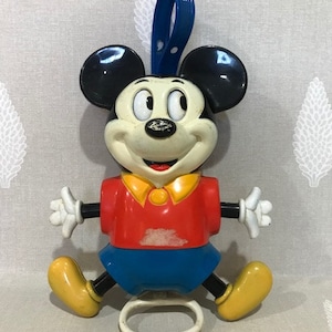May include: A vintage Mickey Mouse toy figure. The figure has black ears, a white face, and a red shirt with blue shorts. The toy has yellow shoes and white gloves. A blue loop is attached to the top of the figure.
