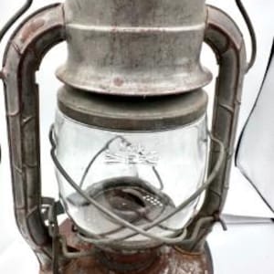 May include: A weathered metal lantern featuring a clear glass globe, a sturdy handle, and a rusted finish. The lantern has a cylindrical top section, a central glass chamber, and a wide base with visible metal texture and signs of age and wear.