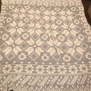 May include: A white, crocheted tablecloth with a detailed floral and geometric pattern. The edges feature a scalloped trim and a repeating design of flowers and diamonds. The intricate openwork creates a delicate, vintage aesthetic.