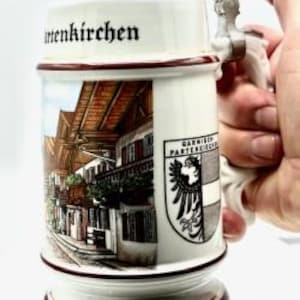 May include: A white ceramic beer stein with a hinged metal lid and handle. The stein features a detailed illustration of a European town scene, the text "Garmisch-Partenkirchen", and a coat of arms. The base and rim are trimmed in burgundy.