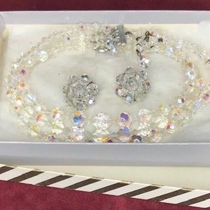 May include: A clear, multi-strand necklace and matching clip-on earrings set. The jewelry is made of iridescent, faceted beads, presented in a white box with a striped border. The necklace has a silver clasp.