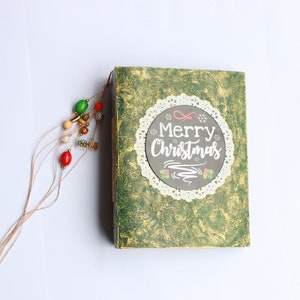 May include: A green journal with a gold and white design. The cover has a white lace circle with the text "Merry Christmas" in white lettering. A brown string with colorful beads is attached to the journal.