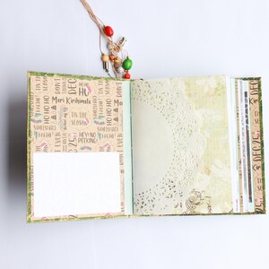 May include: An open journal with a green cover and a brown paper interior. The journal has a white paper insert and a lace doily on the right page. The cover is decorated with Christmas-themed text and images. The text on the cover includes "Meri Kirihimele", "Tis the Season", and "Ho Ho Ho".
