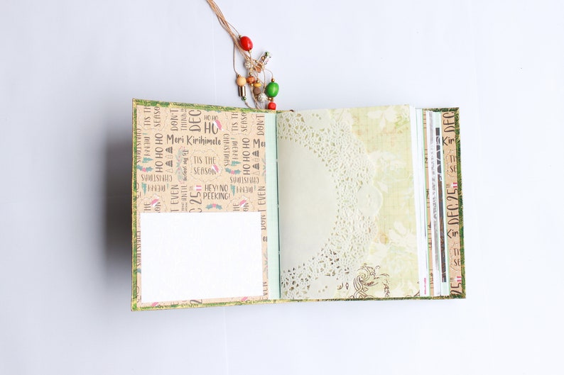 May include: An open journal with a green cover and a brown paper interior. The journal has a white paper insert and a lace doily on the right page. The cover is decorated with Christmas-themed text and images. The text on the cover includes "Meri Kirihimele", "Tis the Season", and "Ho Ho Ho".