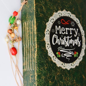 May include: A green journal with gold accents and a white lace doily with the text "Merry Christmas" in black lettering.